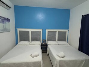 Una habitación con dos camas, cada una con cabecera y ropa de cama blancos. Entre las camas hay una mesita con un florero y un control remoto.