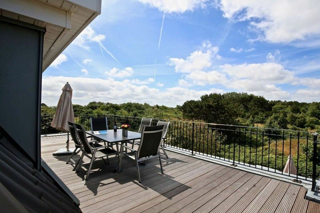 A wooden deck with a table and chairs, a metal railing, and a view of a forested area.