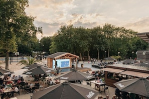 An outdoor seating area with tables and umbrellas, a stage with a screen, and a small building structure.