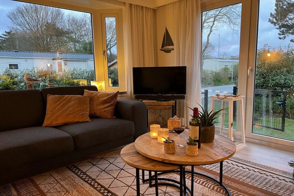 Un salon comprenant un canapé, un téléviseur, une table basse ronde ornée de bougies, et donnant sur un jardin grâce à de grandes fenêtres.