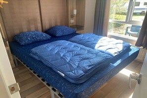Una cama con un edredón azul y almohadas, una mesita de noche con una lámpara y una ventana con vistas al exterior.