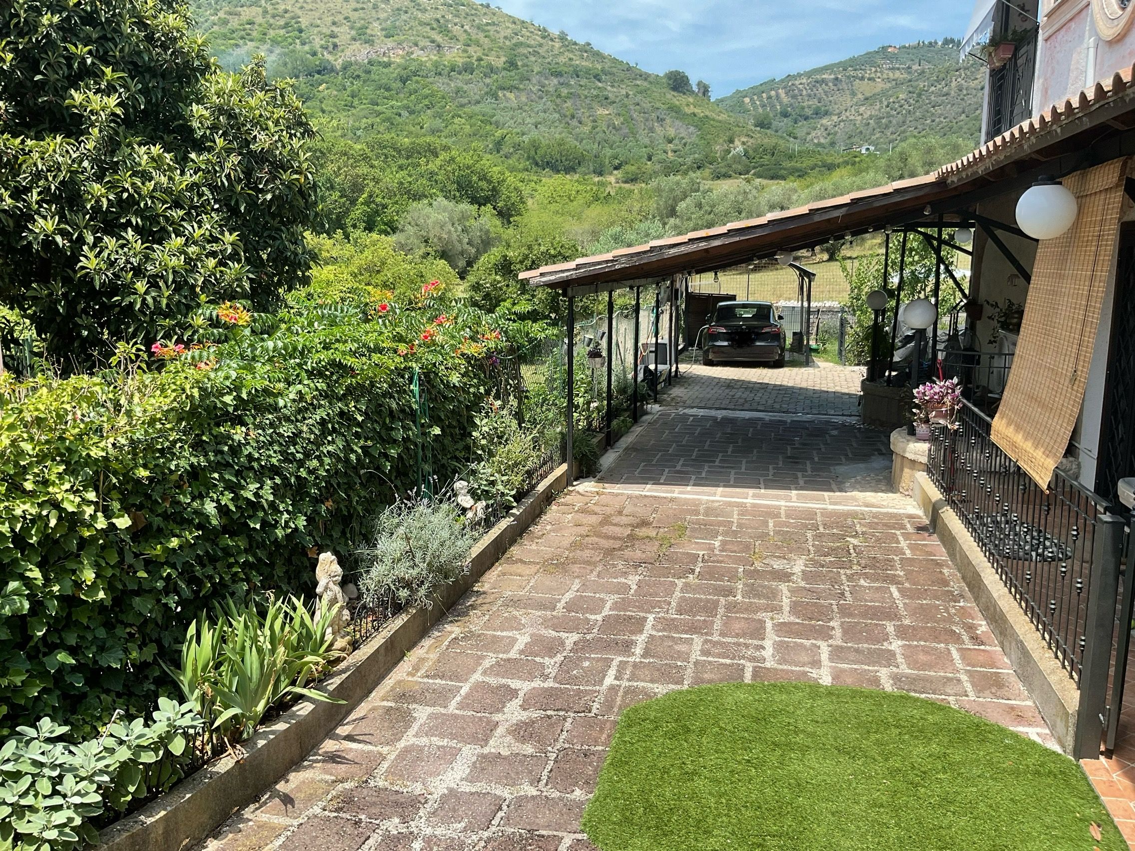 Ein gepflasterter Weg führt zu einem überdachten Parkplatz mit Blick auf eine üppige grüne Landschaft und Hügel im Hintergrund.