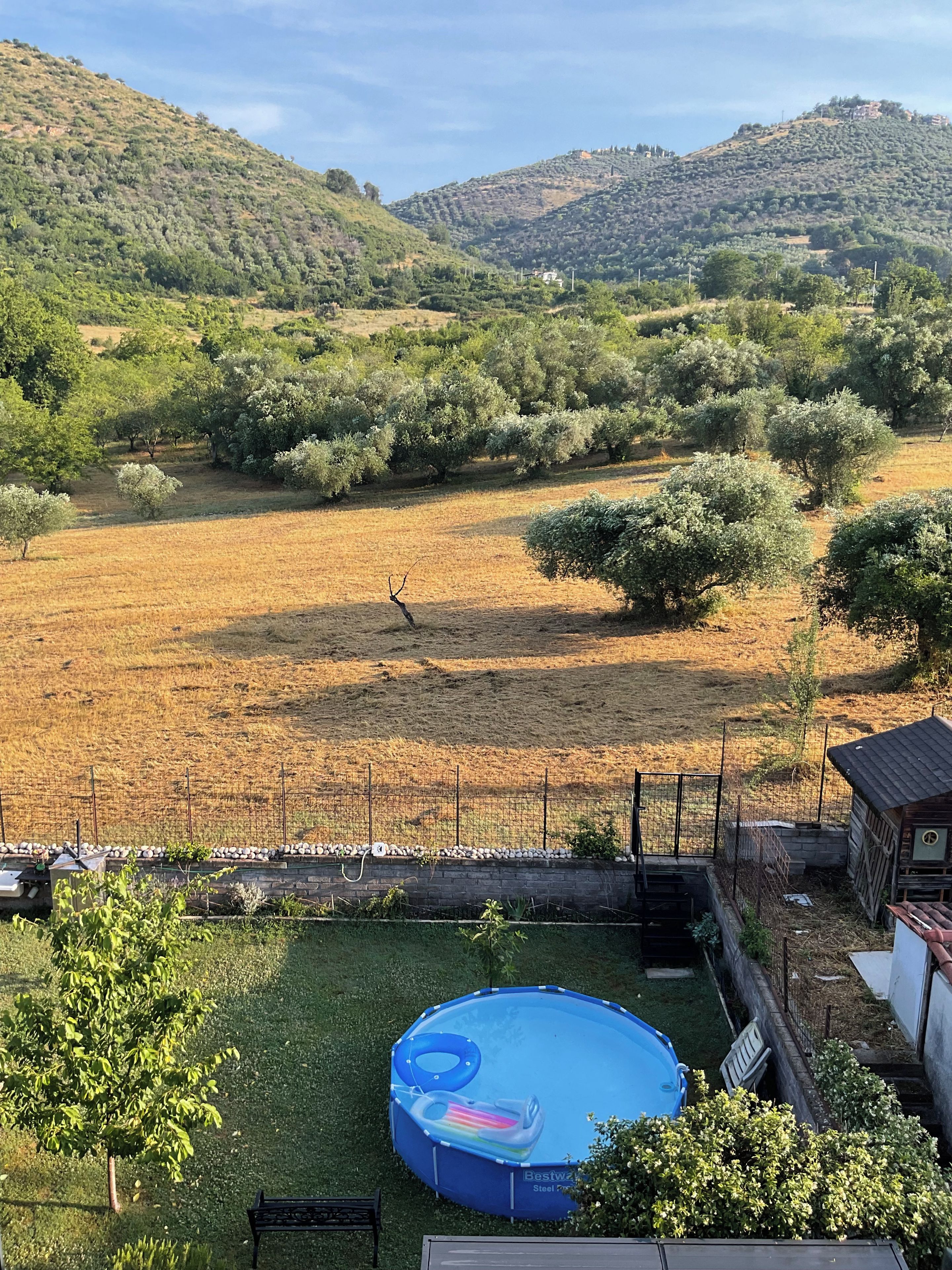 Ein Garten mit einem blauen aufblasbaren Pool, einem eingezäunten Bereich und Blick auf eine hügelige Landschaft.