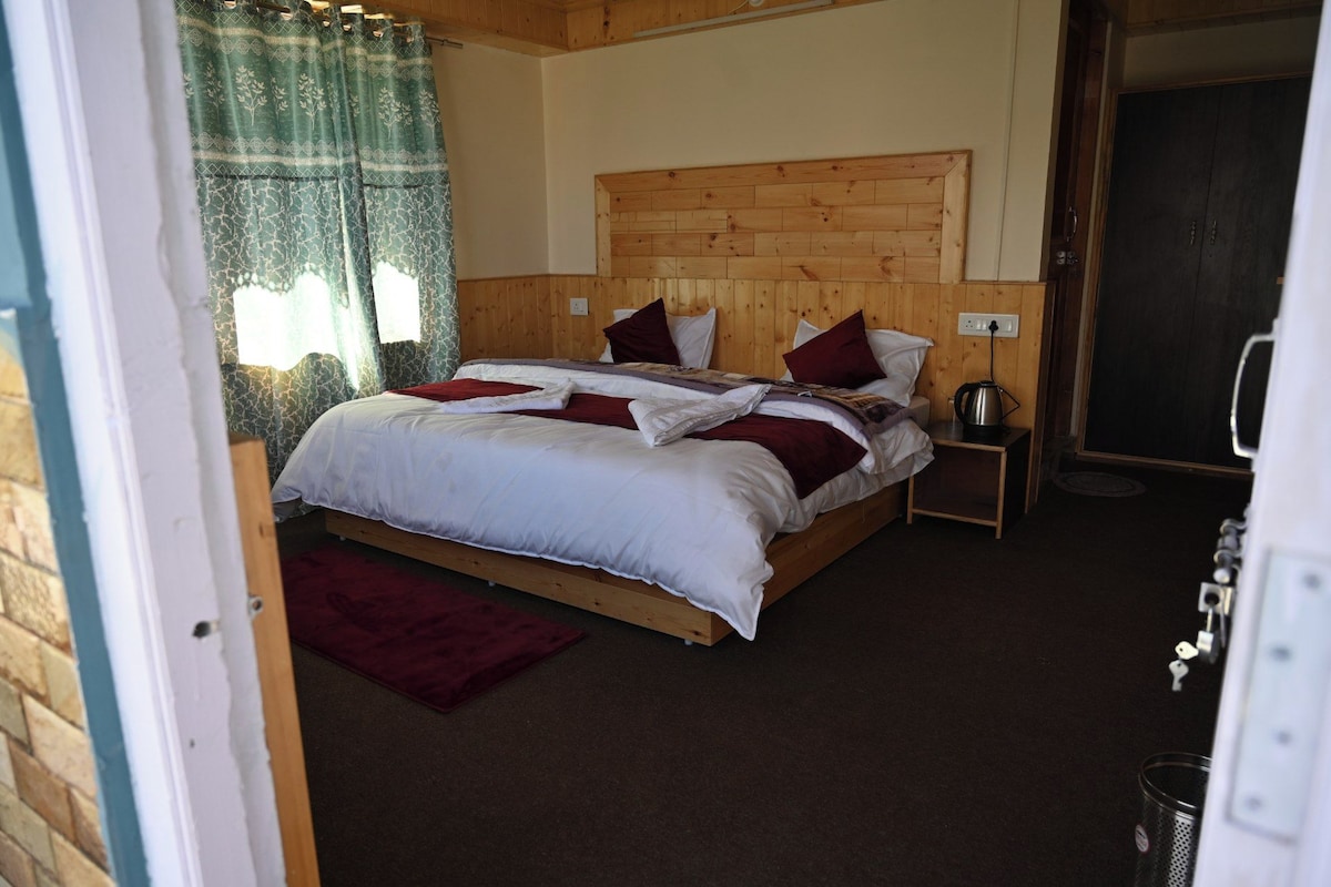 A bedroom with a wooden headboard, a bed with white bedding and red accents, a nightstand with a kettle, and a window with patterned curtains.