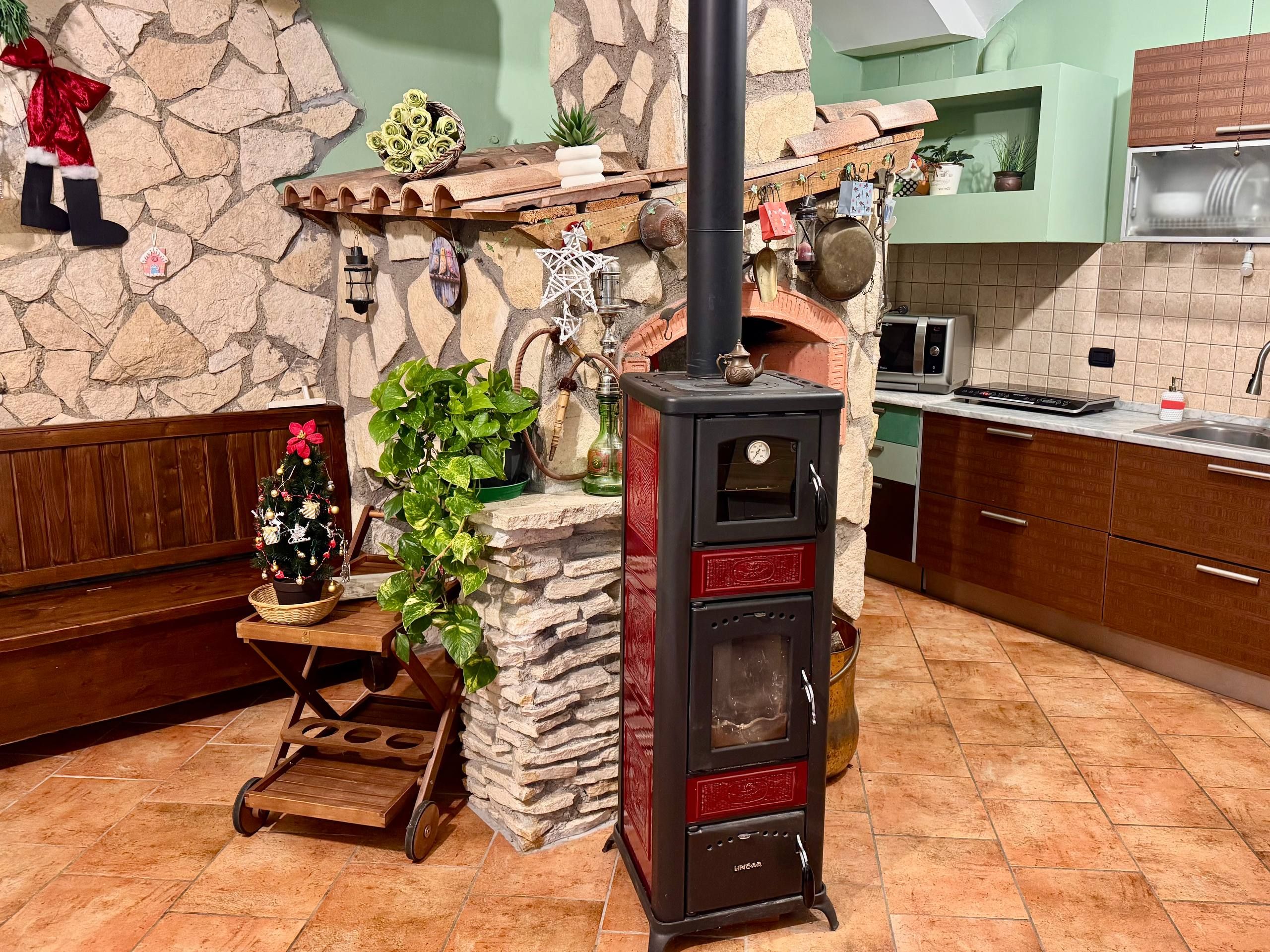 A kitchen with a stone wall, a wood-burning stove, a wooden cart, and a Christmas tree.
