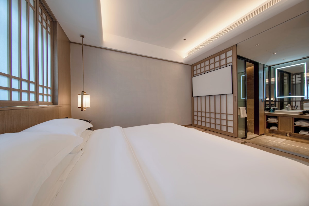 Een hotelkamer met een bed, nachtkastje, hanglamp, een schuifdeur naar een badkamer en een grote spiegel.