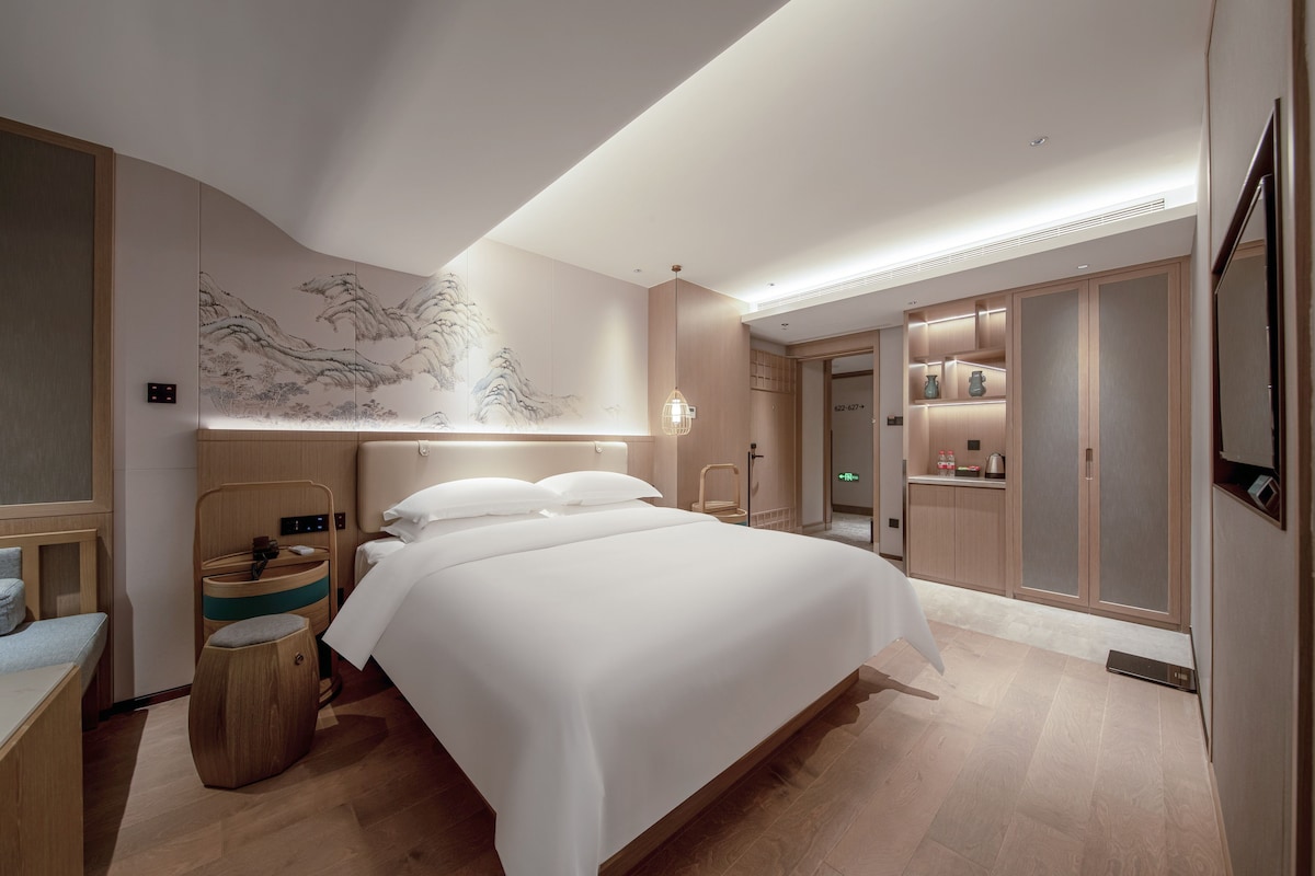 Een moderne hotelkamer met een groot bed, een houten hoofdbord, een muur met een klassiek schilderij, een klein houten krukje en een inbouwkast met spiegel.
