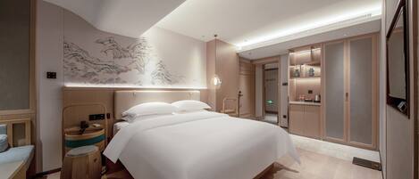 A modern hotel room with a large bed, wooden headboard, a wall with a traditional painting, a small wooden stool, and a built-in wardrobe with a mirror.