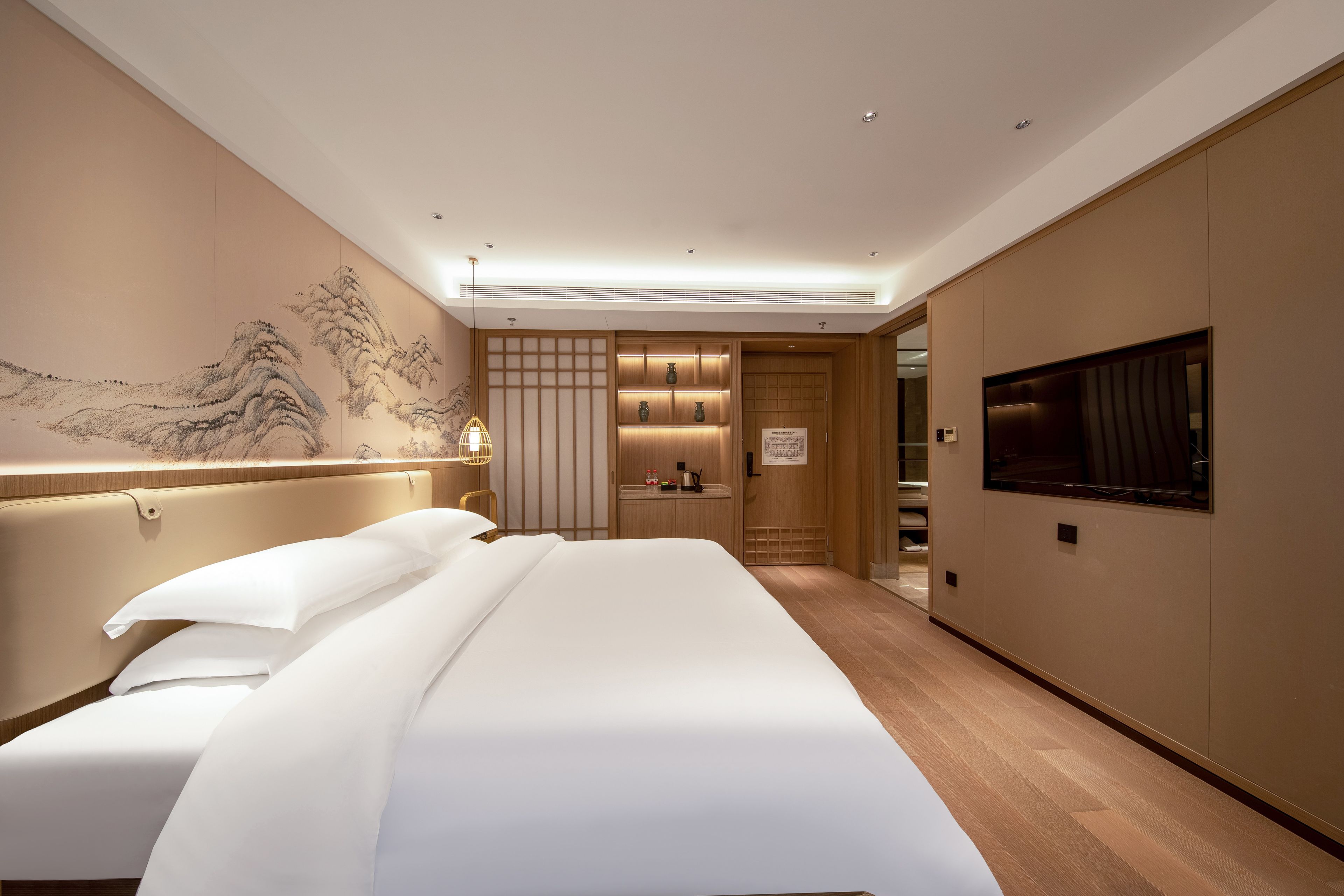 Une chambre d’hôtel avec un grand lit, une télévision, un mur orné d’un tableau traditionnel et une porte coulissante donnant sur une salle de bain.