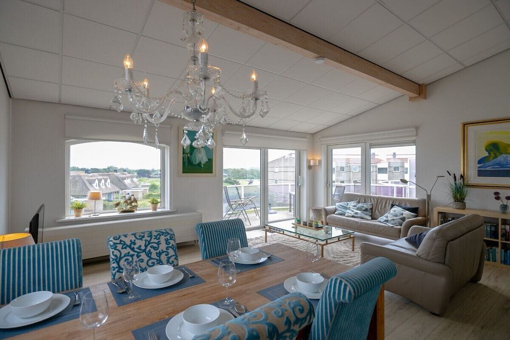 A spacious living room with a dining area, large windows, a chandelier, and a view of the city.