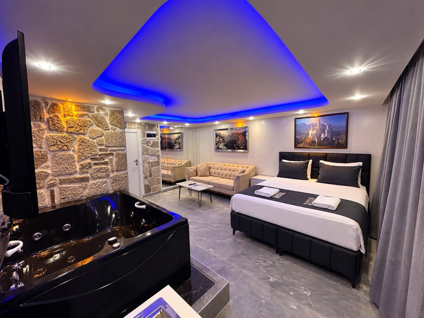 A modern hotel room with a large bed, a sofa, a jacuzzi, and a stone wall.