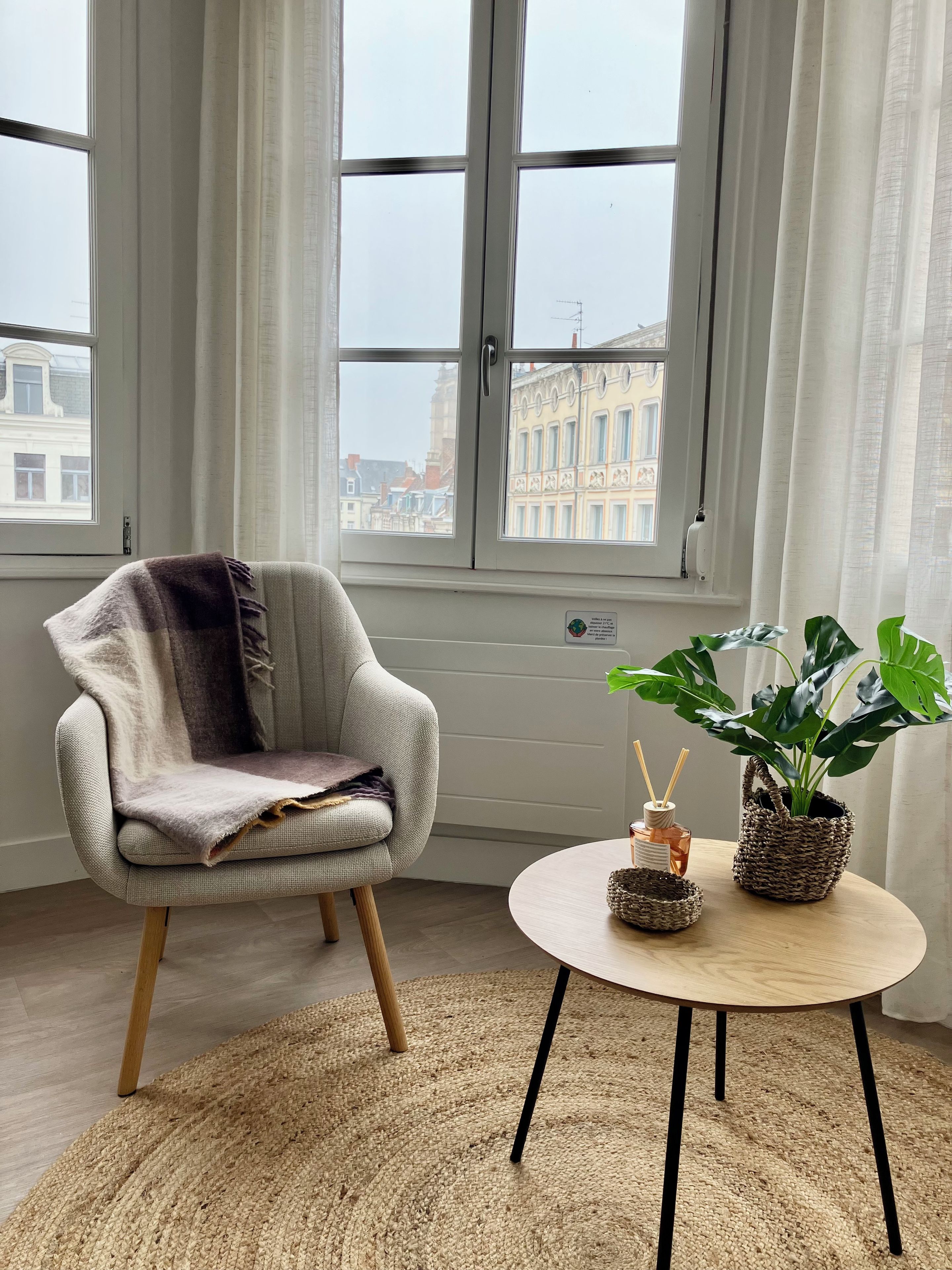 Een kamer met een raam, een stoel, een ronde tafel met een plant en een klein decoratief object.
