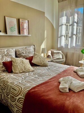 A neatly arranged bedroom with a patterned bedspread, multiple pillows, a bedside table with a lamp, and artwork on the wall. - New Listing (Douai)