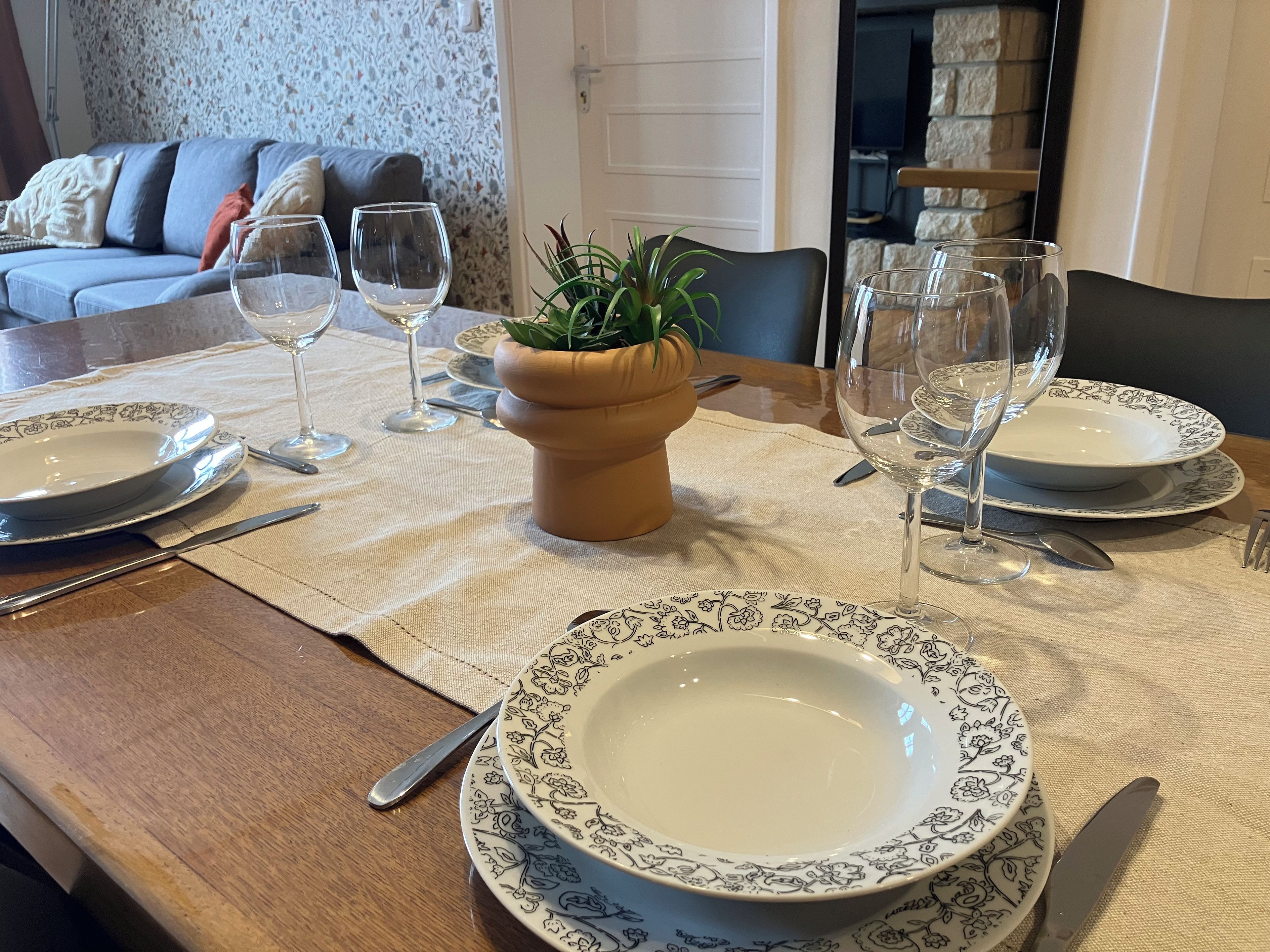 Un tavolo da pranzo apparecchiato con piatti, bicchieri e posate, un vaso con una pianta e un divano sullo sfondo.