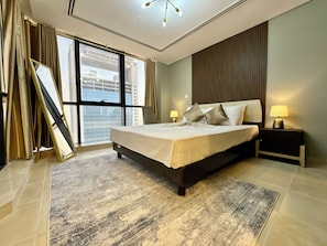 A hotel room with a large bed, a nightstand, a lamp, a window with curtains, and a view of buildings outside. - Cosmos Living Signature Studio in Al Furjan (Dubai)