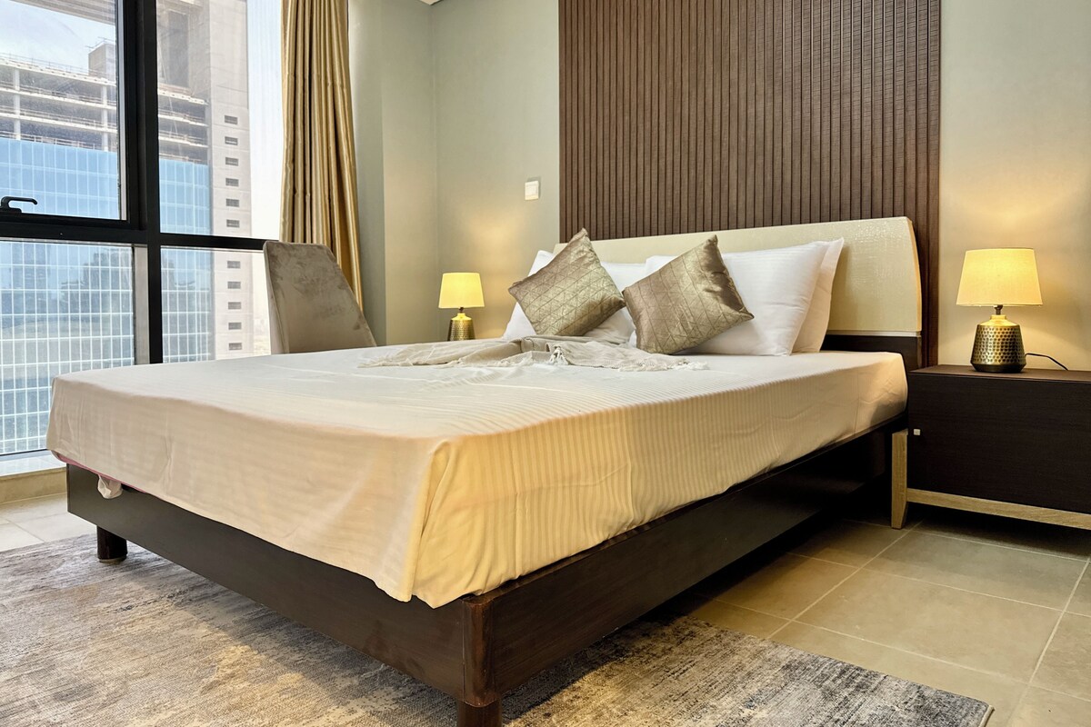 A hotel room with a large bed, two bedside lamps, a view of the city through a window, and a wooden panel wall.