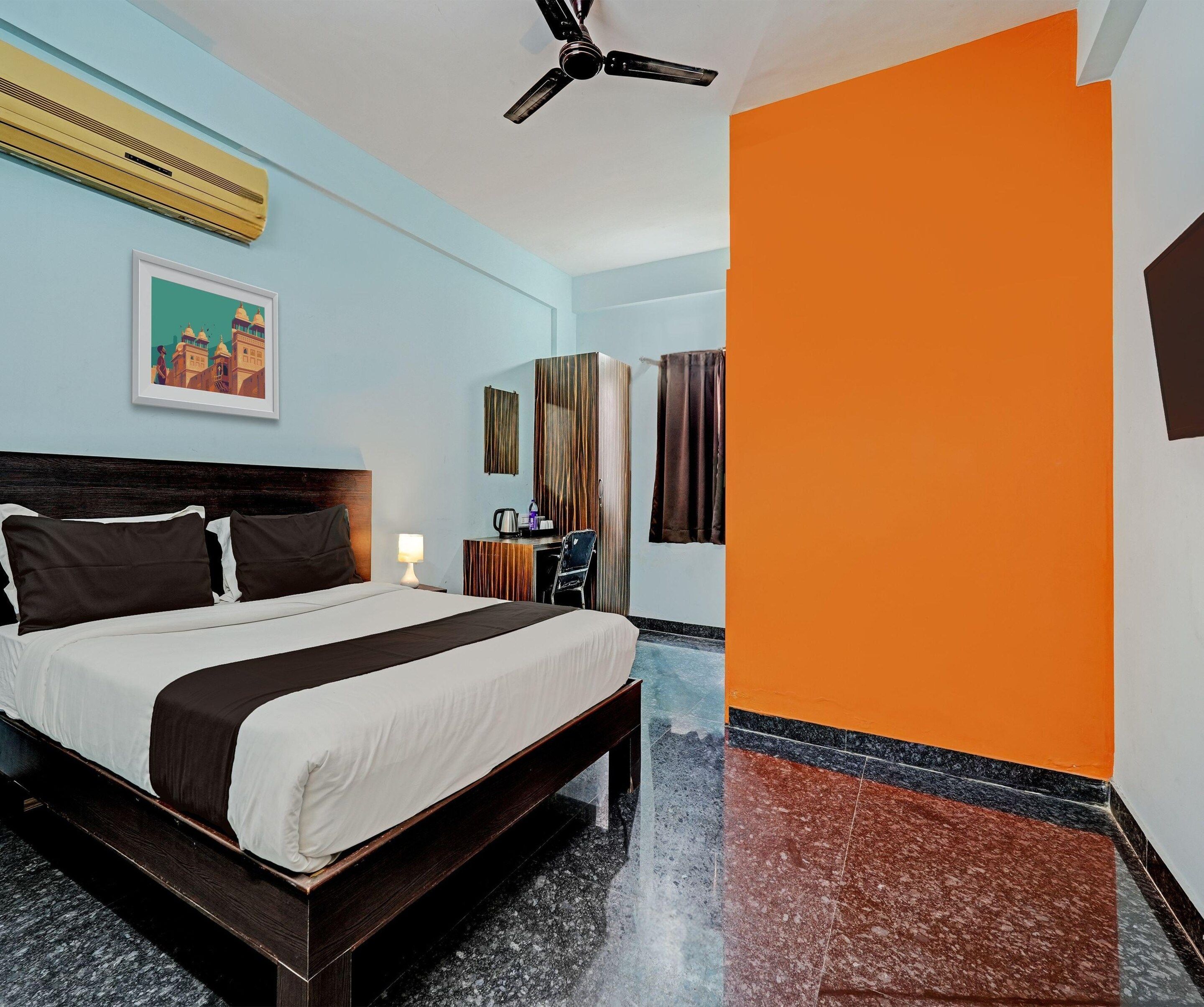 Une chambre d’hôtel avec un lit, un bureau, une chaise, un ventilateur de plafond et un tableau au mur.