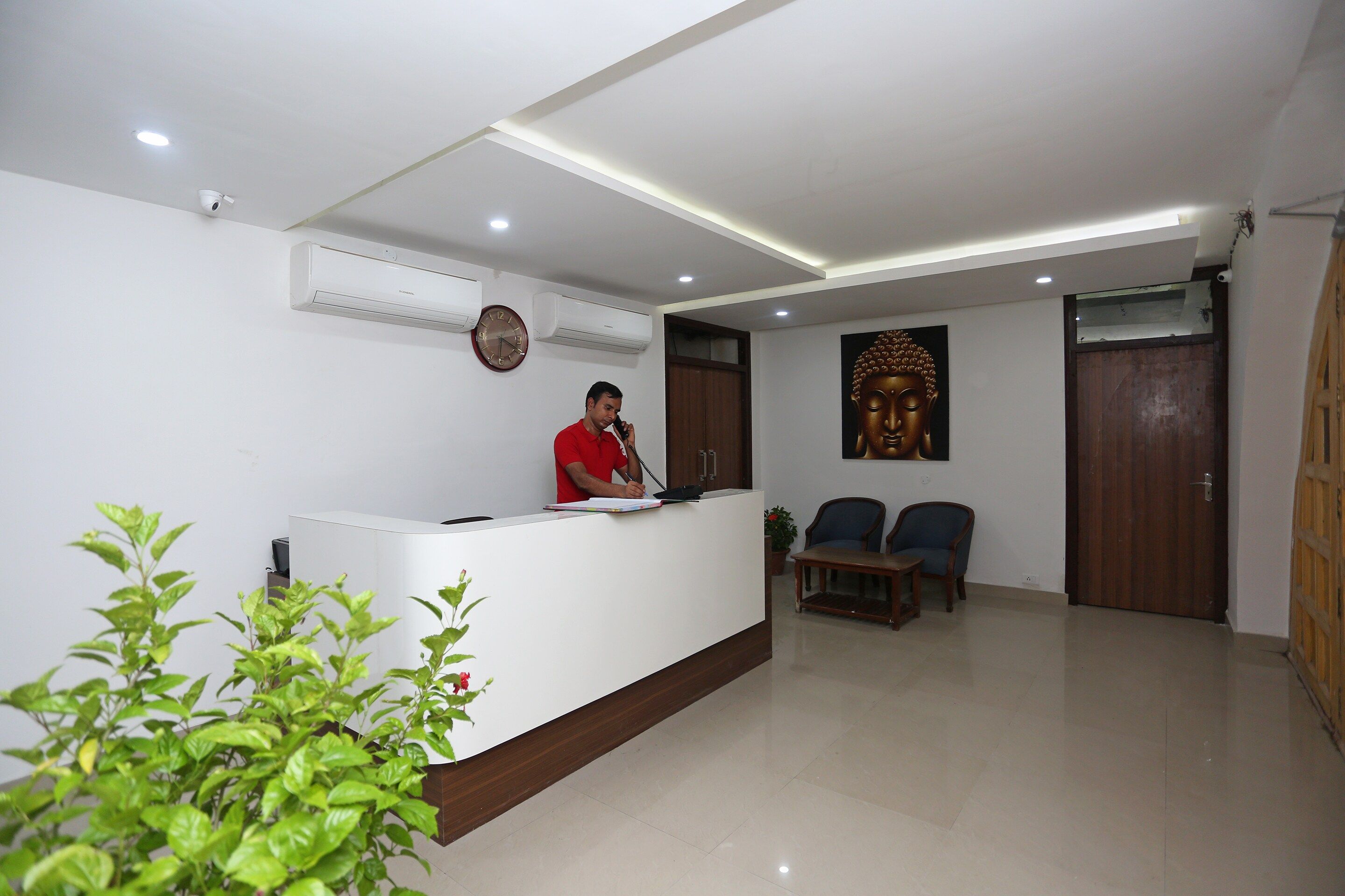 A reception area with a receptionist, a clock, an air conditioning unit, a Buddha statue, and a wooden door.