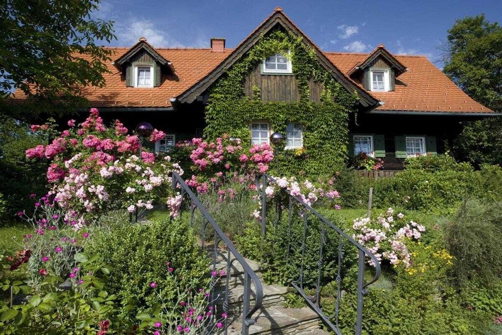 Ein zweigeschossiges Haus mit rotem Ziegeldach, efeuüberwucherten Mauern und einem Garten mit rosa Blumen.