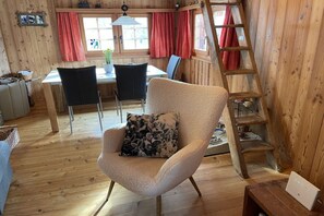 A cozy cabin interior with a wooden floor, a dining area with a table and chairs, a cushioned armchair with a floral pillow, and a wooden ladder.