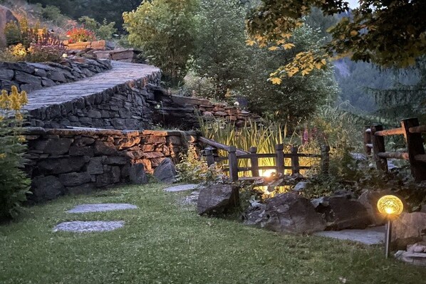 A stone pathway leading to a wooden bridge over a small stream, surrounded by lush greenery and a stone wall.