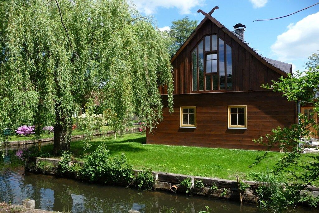 A wooden house with a sloped roof, large windows, and a pond in the foreground.