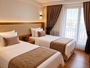 Quarto de hotel com duas camas, cabeceira de madeira, mesa de cabeceira com telefone e janela com cortinas.