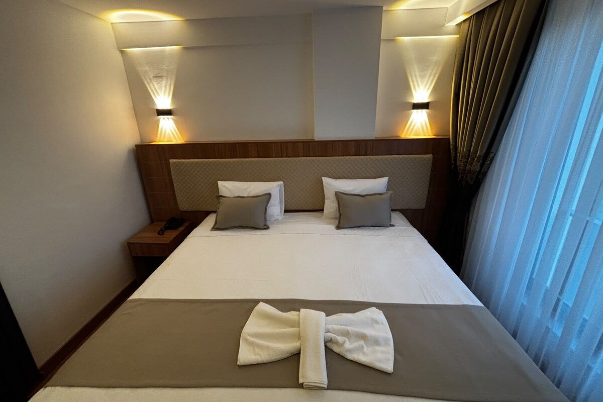 A hotel room with a bed, two bedside lamps, a nightstand, and a window with curtains.