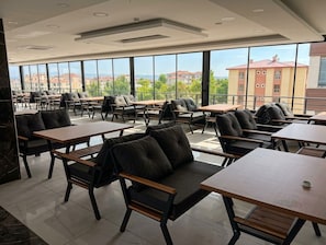 Een ruime binnenruimte met meerdere tafels en stoelen, grote ramen met uitzicht op gebouwen en groen.