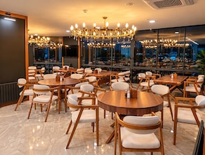 Restaurante com várias mesas e cadeiras redondas, amplas janelas e um lustre.
