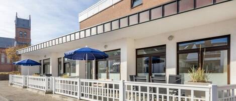 A building with a white fence, outdoor seating with blue umbrellas, and a brick facade.