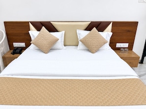 A neatly made bed with a quilted headboard, two bedside tables with a phone and glasses, and a bedside lamp. - Hotel Shreedha Palace (Indore)