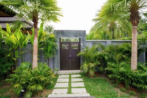 A gated entrance with a pathway leading to a building, surrounded by lush greenery and palm trees.