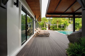 A wooden deck with lounge chairs, a swimming pool, and a view of tropical vegetation.