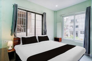Une chambre d’hôtel avec un grand lit, deux tables de chevet, une petite table, une lampe et deux fenêtres avec des rideaux.