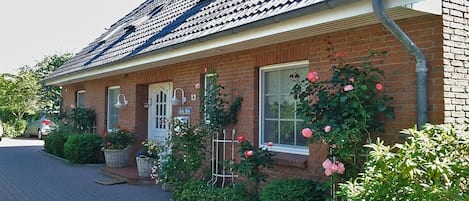 A brick house with a tiled roof, a front porch with a white door, and a garden with pink roses.