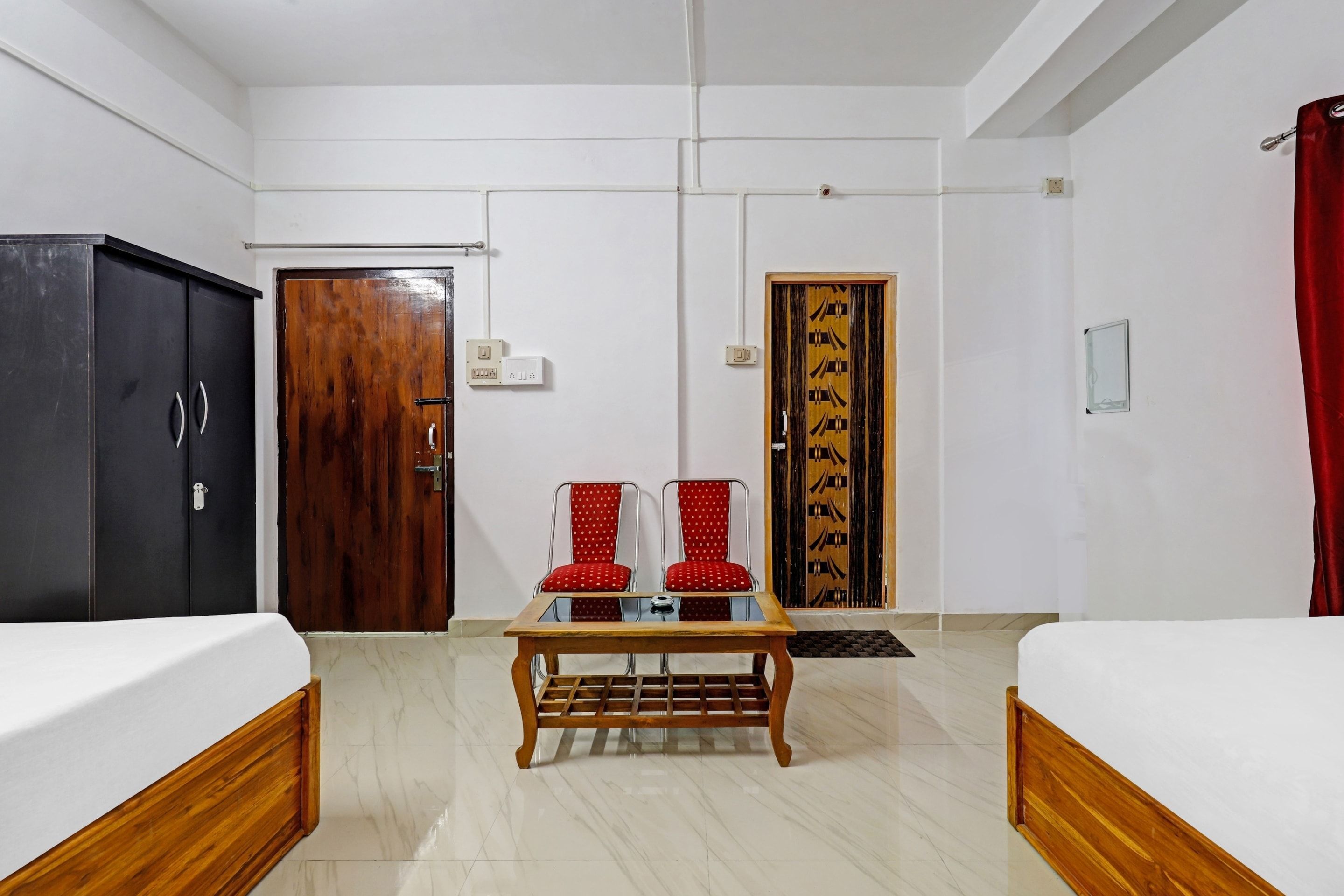 Habitación de hotel con dos camas, una mesa de madera, dos sillas rojas, un armario y una puerta con un panel decorativo.