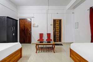 Habitación de hotel con dos camas, una mesa de madera, dos sillas rojas, un armario y una puerta con un panel decorativo.