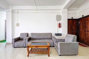 A modern living room with a grey sofa, a wooden coffee table, and a grey armchair. There is a small table with a black electronic device, a fire extinguisher mounted on the wall, and a wooden door with a number. - Hotel O Assam Inn (Guwahati)