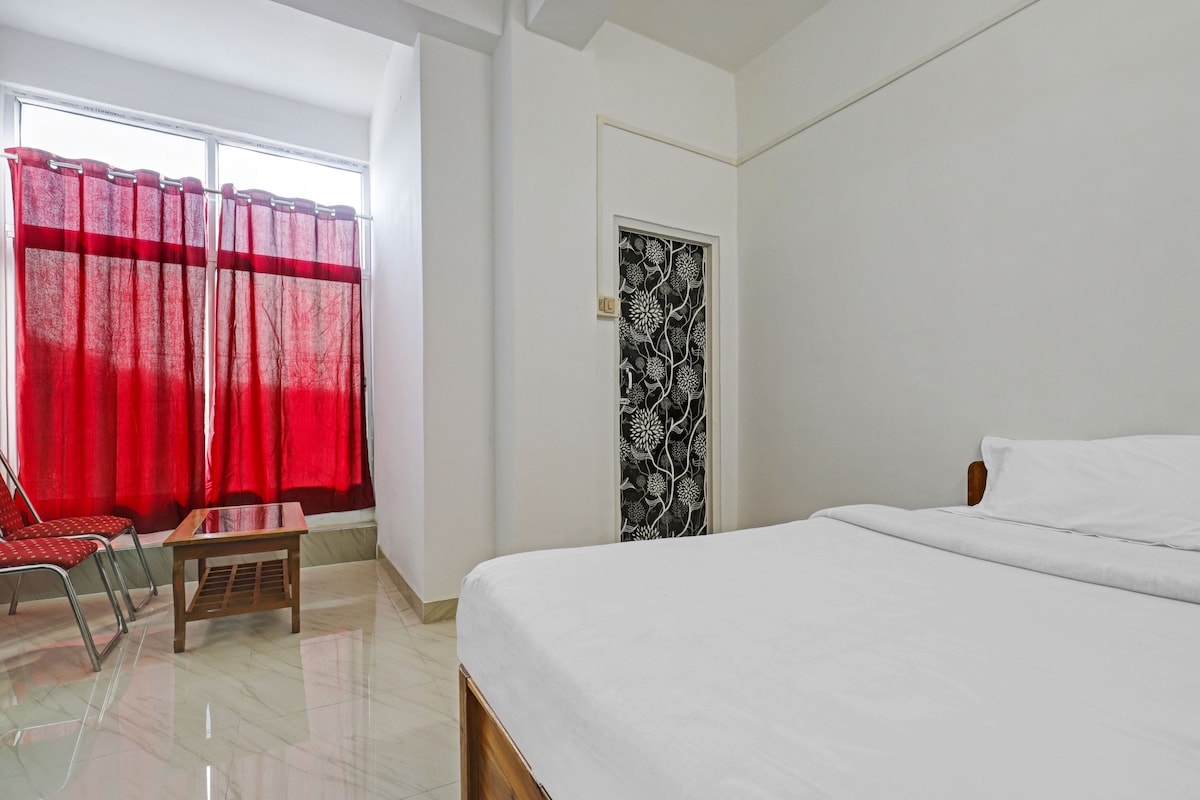 A hotel room with a bed, a chair, a small table, a window with red curtains, and a door with a black and white pattern.