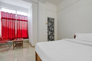 A hotel room with a bed, a chair, a small table, a window with red curtains, and a door with a black and white pattern. - Hotel O Assam Inn (Guwahati)