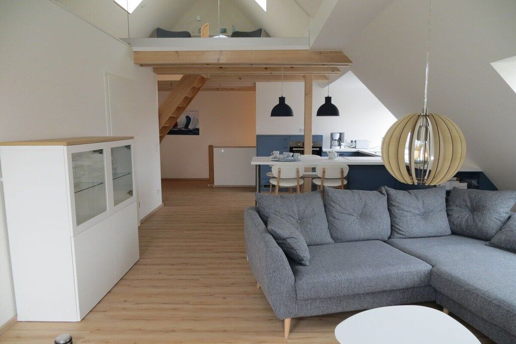 A modern loft-style living space with a wooden floor, a grey sectional sofa, a dining area with a white table and chairs, and a kitchen in the background.