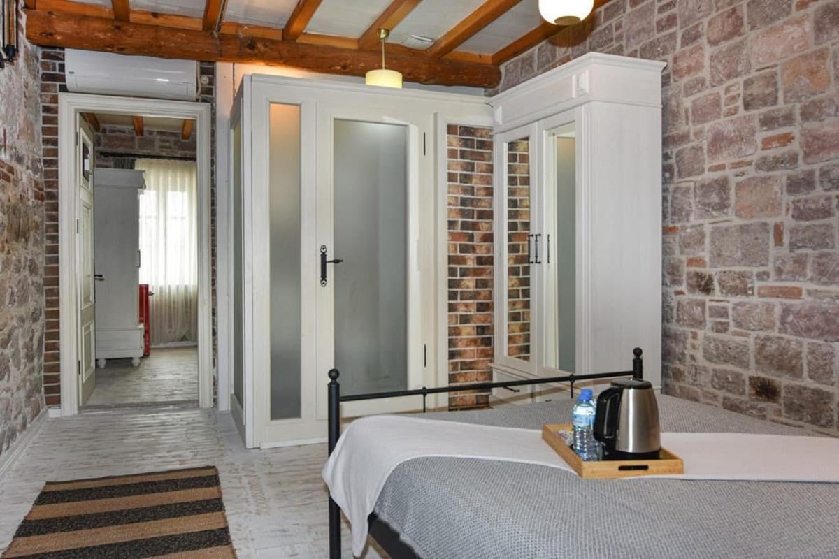Une pièce comprenant un lit, un banc en bois avec une bouilloire et une bouteille d’eau, une armoire blanche et un mur en briques.
