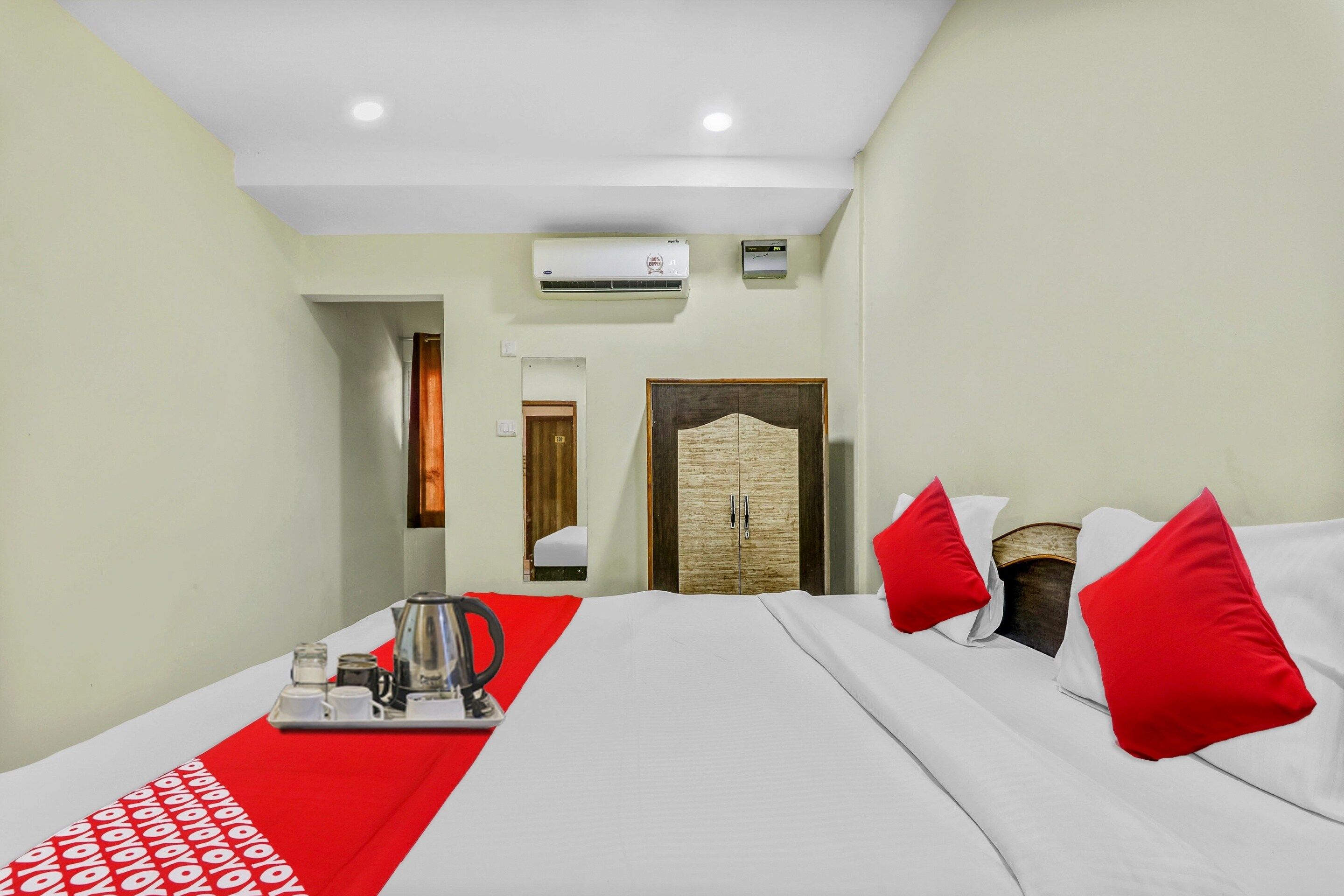 A hotel room with a bed, a coffee table with a red runner, a wall-mounted air conditioner, and a door with a mirror.