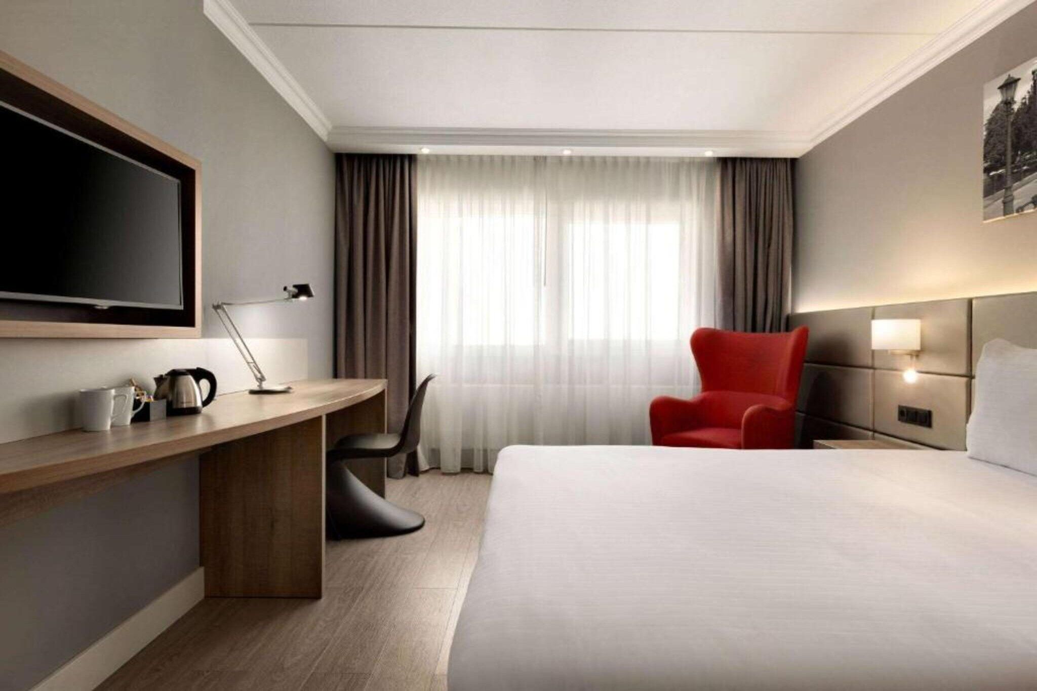 Una camera d'albergo con un letto, una scrivania con una lampada, una sedia, una televisione e una finestra con tende.