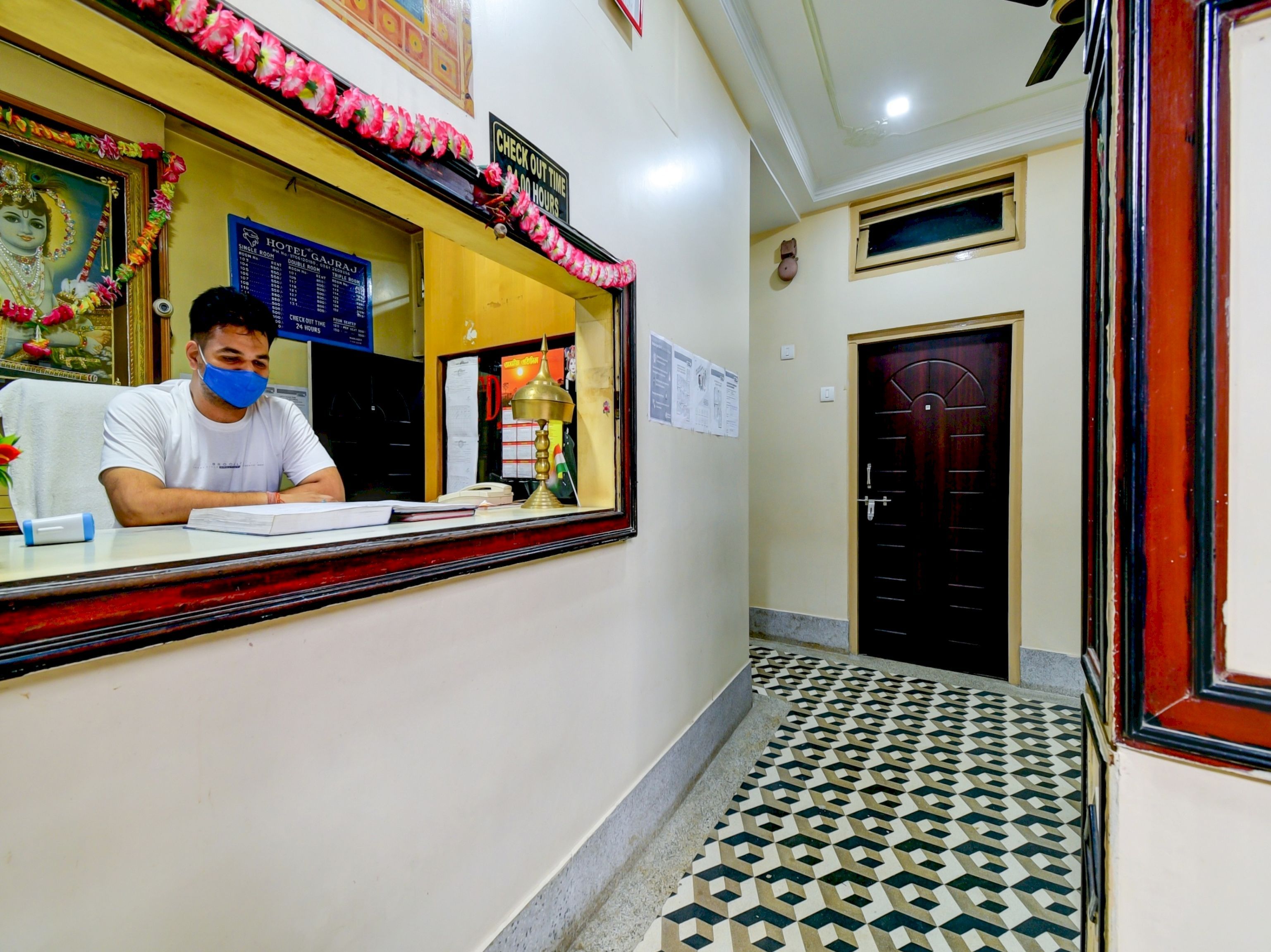 A hotel reception desk with a man seated behind it, a check-out time sign, and a framed picture above the desk.
