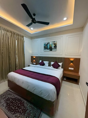 A bedroom with a bed, a ceiling fan, a nightstand, a rug, and a wall-mounted light fixture. - VM Anandam (Varanasi)