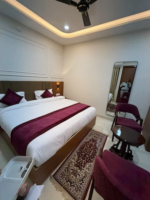 A hotel room with a bed, a desk, a chair, a ceiling fan, and a mirror. - VM Anandam (Varanasi)