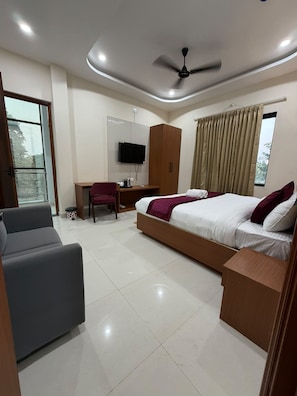 A modern hotel room with a bed, a desk, a chair, a television, and a ceiling fan. - VM Anandam (Varanasi)