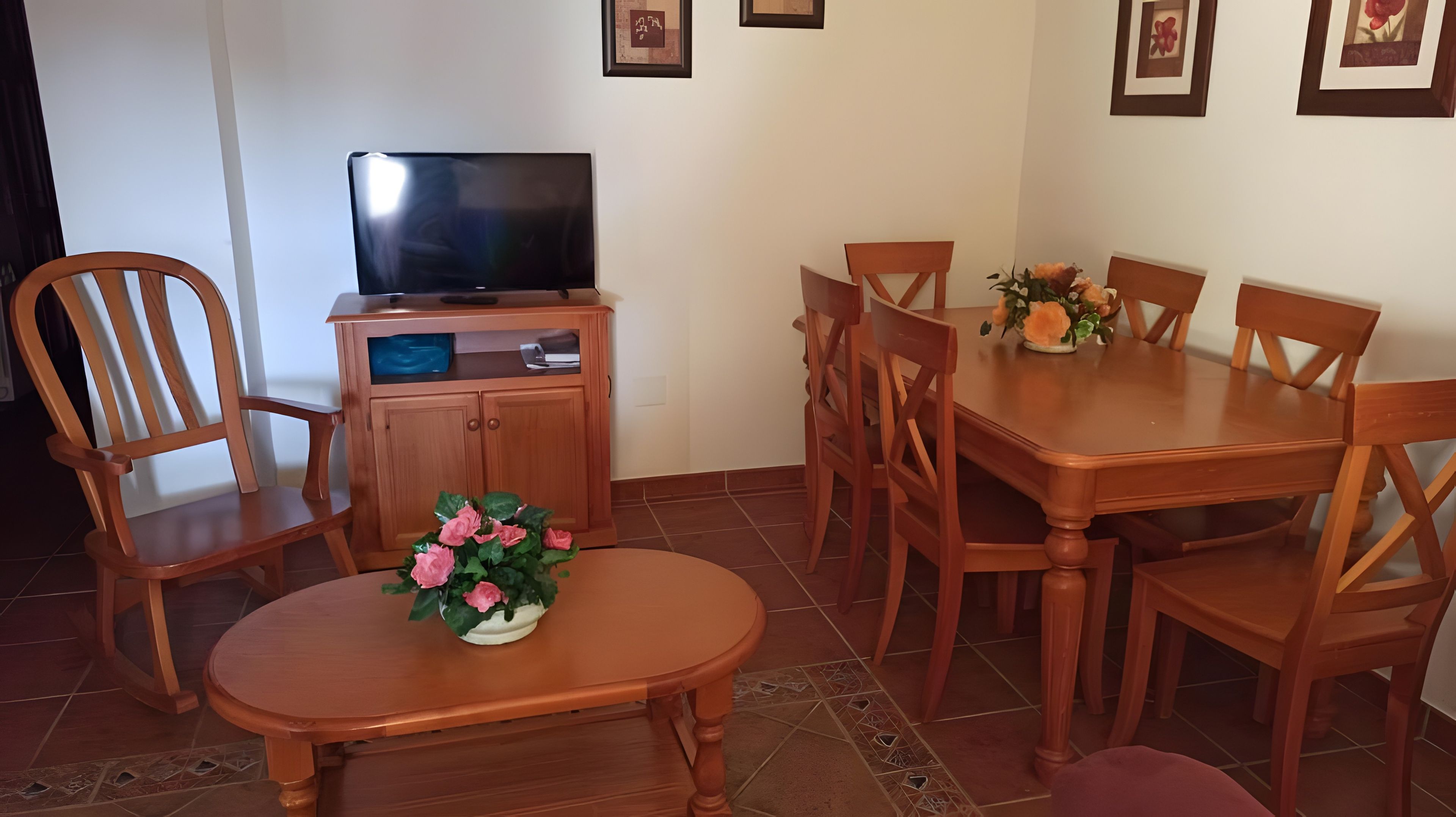 Un comedor con una mesa de madera, sillas, un televisor en un mueble y un jarrón de flores.